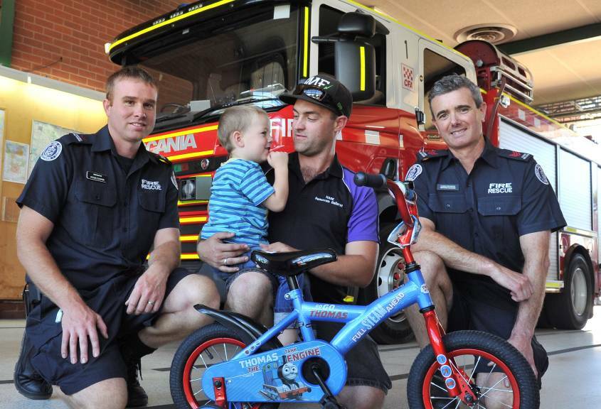 FIERY WHEELS: Firefighters Peter Cooper (left) and Nathan Duggan (right) give Keith Bassett, 3, and his dad Courtney a new bike. Picture: Lachlan Bence