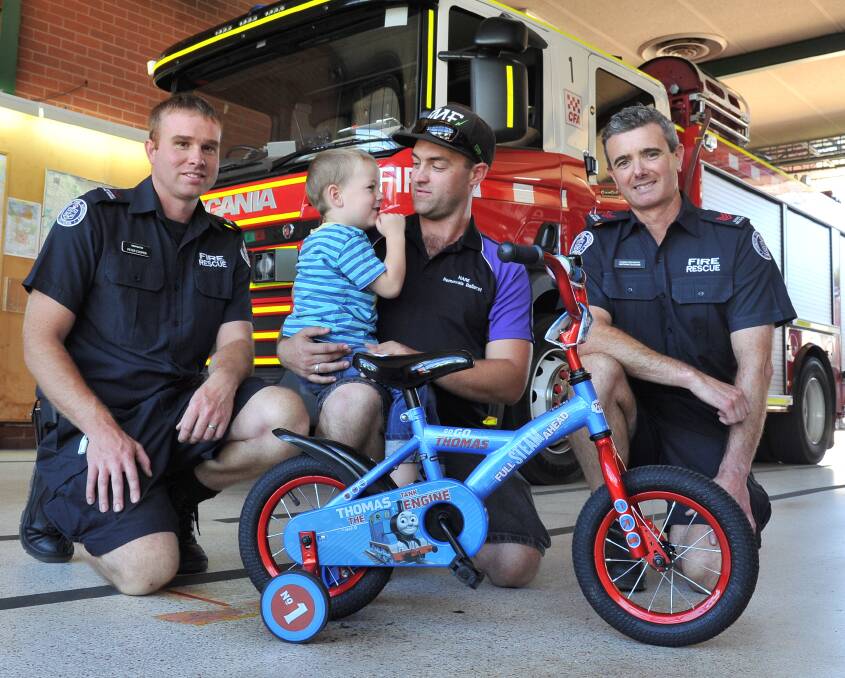 FIERY WHEELS: Firefighters Peter Cooper (left) and Nathan Duggan (right) give Keith Bassett, 3, and his dad Courtney a new bike. Picture: Lachlan Bence 